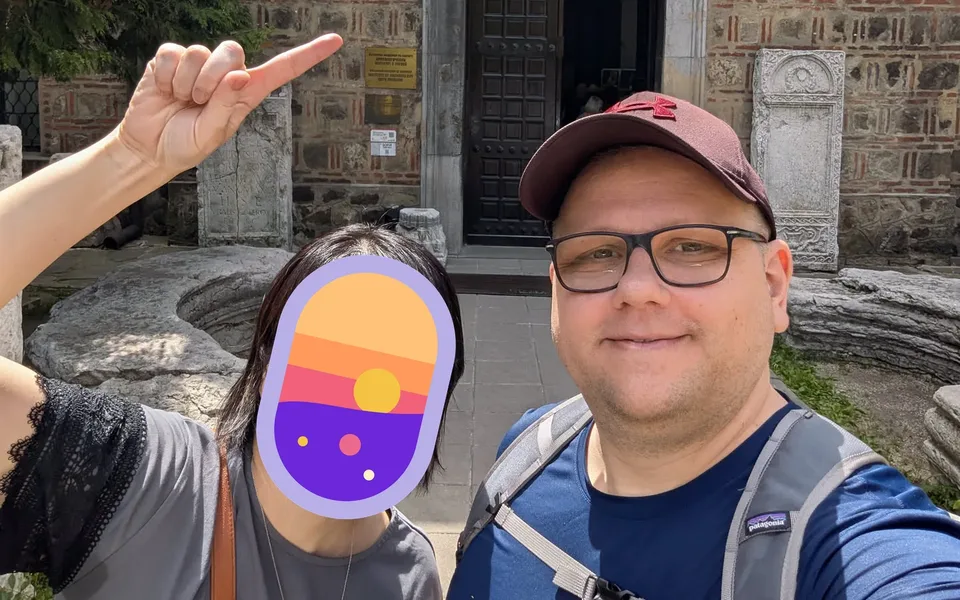 Wolfi's wife pointing up in front of a museum, face covered by a littledots icon, with Wolfi smiling beside